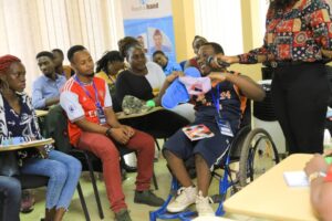 A group of individuals in a classroom setting, with a person in a wheelchair demonstrating an item while others observe attentively.