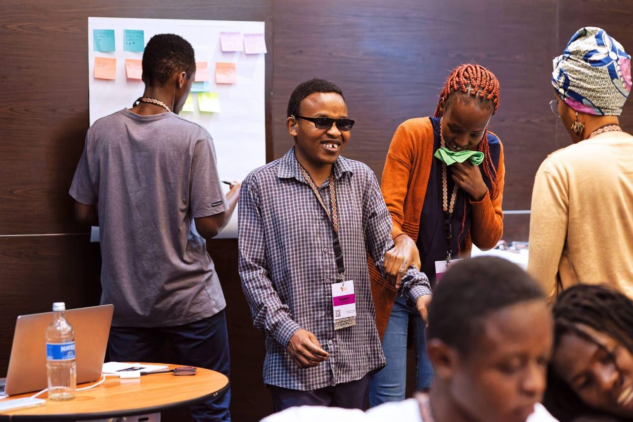 Group of individuals engaged in a collaborative activity, with one person with a visual impairment wearing sunglasses and smiling, surrounded by sticky notes and laptops.