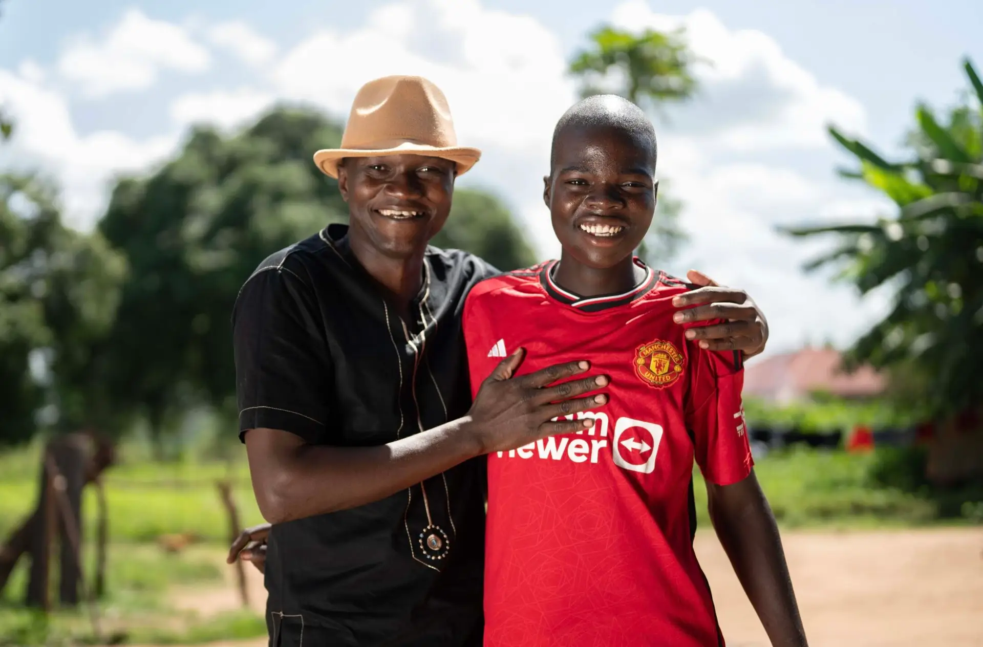 aria-label | A young man with a hat proudly supporting a young boy in a Manchester United football shirt. They are smiling into the camera.