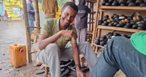 A young Ethiopian man sits on a stool in his shop smiling and shining another man's shoes
