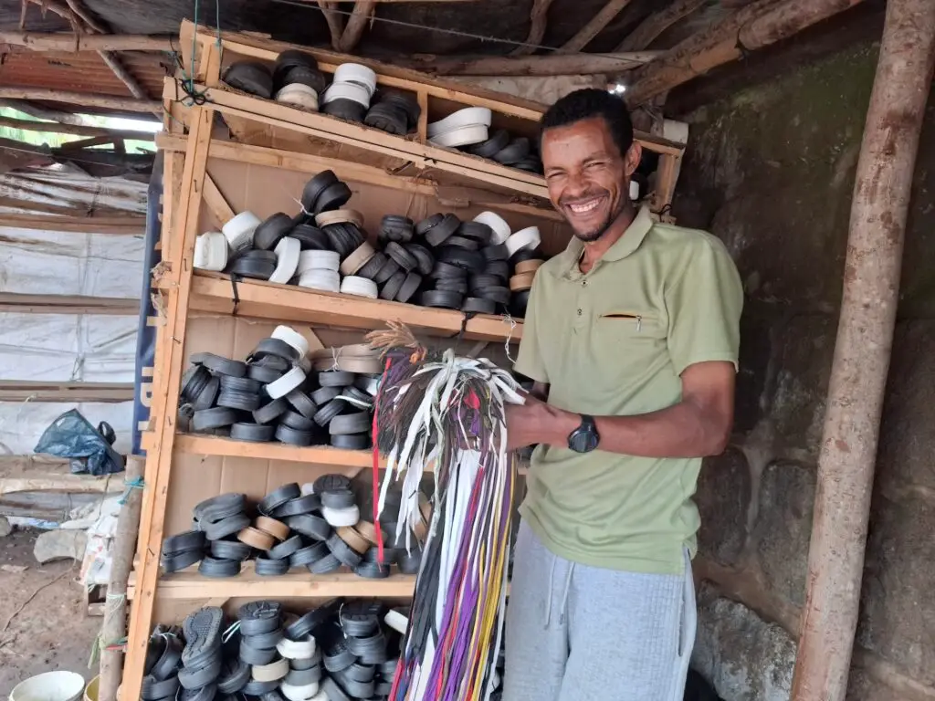 A young Ethiopian man in a green shirt stands smiling holding shoelaces in front of a shelf filled with shoes