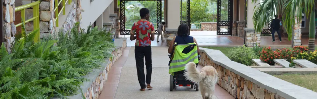Ashesi campus walkway with lush greenery, a person in a wheelchair wearing a high-visibility vest, and a companion in a red floral shirt near stone walls.