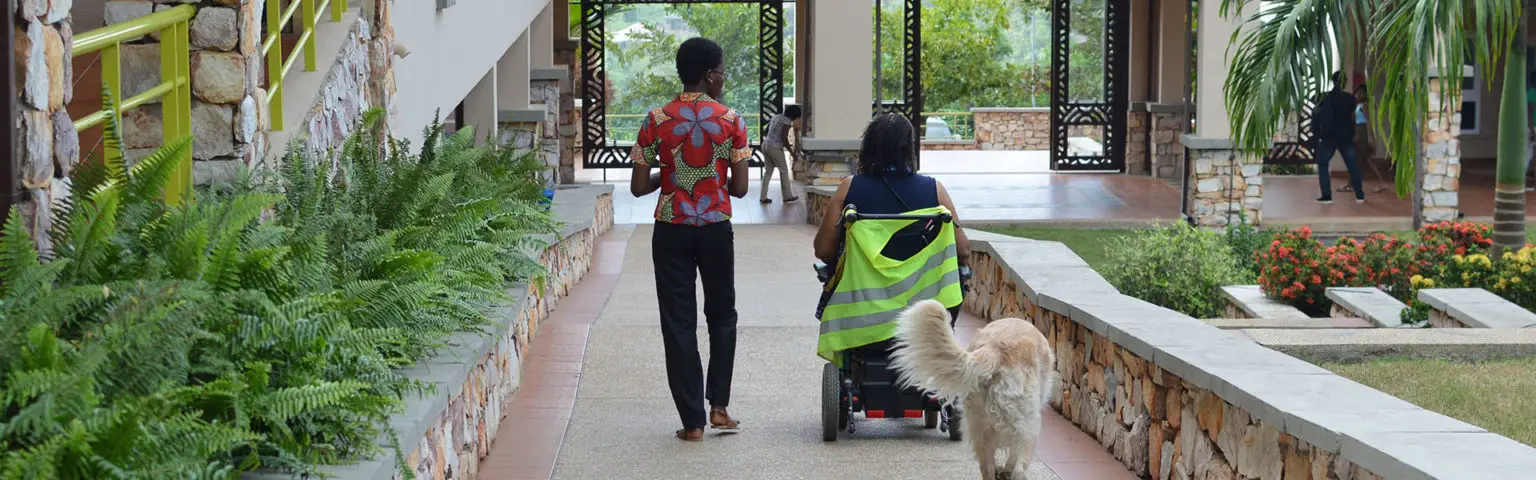 Ashesi campus walkway with lush greenery, a person in a wheelchair wearing a high-visibility vest, and a companion in a red floral shirt near stone walls.