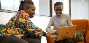 A woman in a brightly colored Ankara print jacket helps a young woman in a grey sweater on a laptop. They are both smiling.