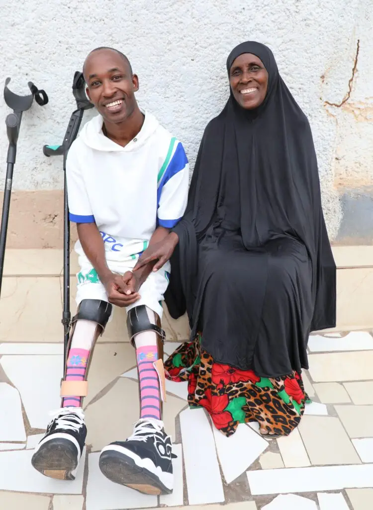 A young man with crutches sits smiling next to an older woman wearing a hijab