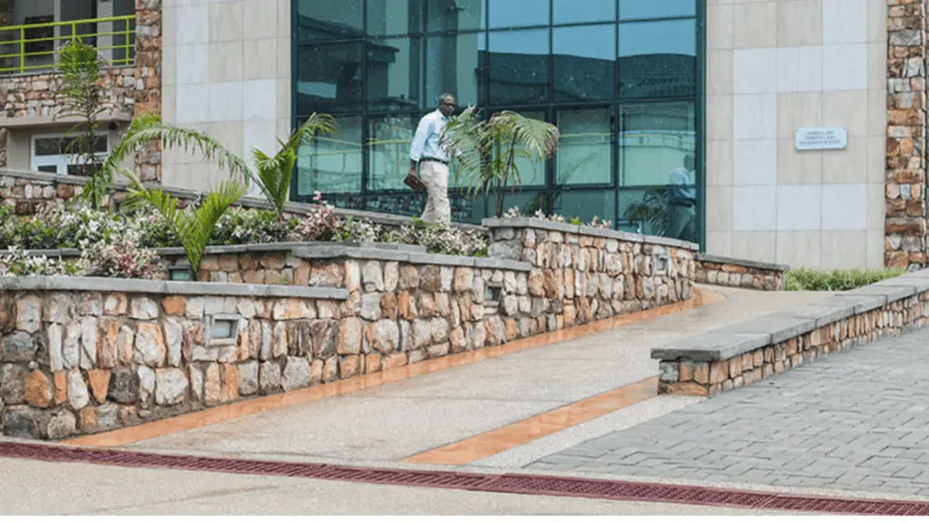 Ashesi campus exterior with a glass facade, stone retaining walls, and tropical plants; a man in light shirt and beige pants walks along the walkway and wheelchair ramp.