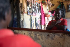 A young woman in a red shirt looks on the mirror and signs the Ugandan Sign Language sign for love