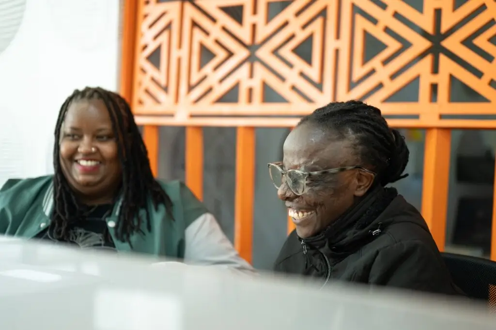 Two young women sit at a desk in an open floor plan office talking and laughing together