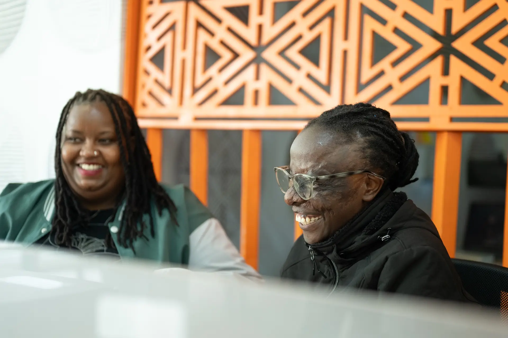 Two young women sit at a desk in an open floor plan office talking and laughing together