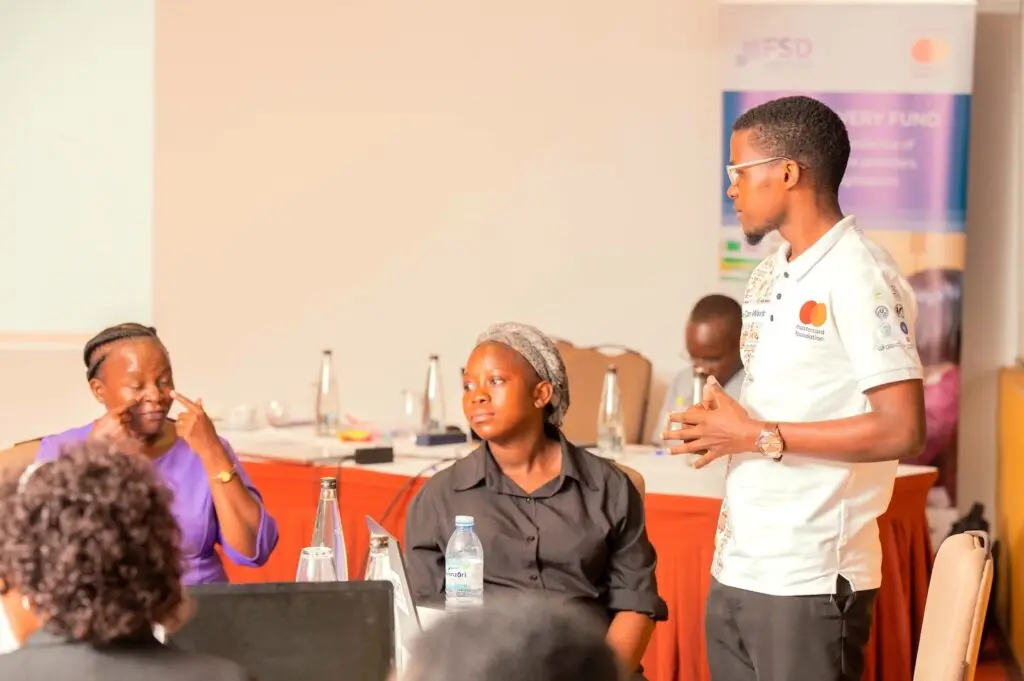 A man with glasses and a yellow We Can Work shirt stands in front of a room of people watching the sign language interpreter
