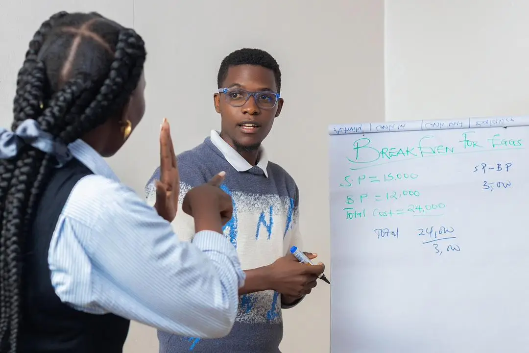 A man with glasses writes on a flip chart while having a conversation with a woman in braids using sign language